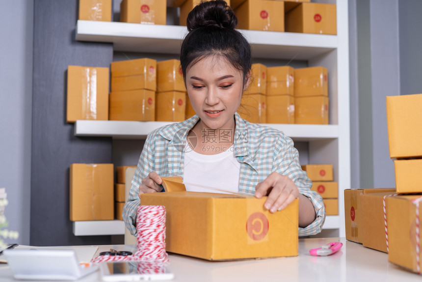 网上女青年创业者使用磁带在家庭办公室包装裹箱准备产品提供给客户图片