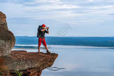 背着包的男旅行者使用相机拍摄照片在悬崖边缘岩石山顶高清图片