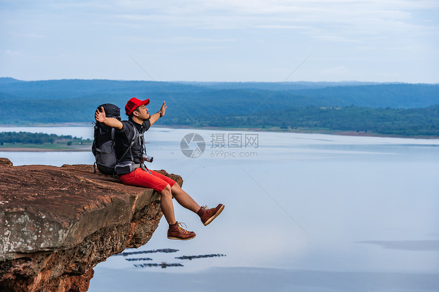 欢乐的徒步男子坐着和手势举起臂在悬崖边缘岩石山顶图片
