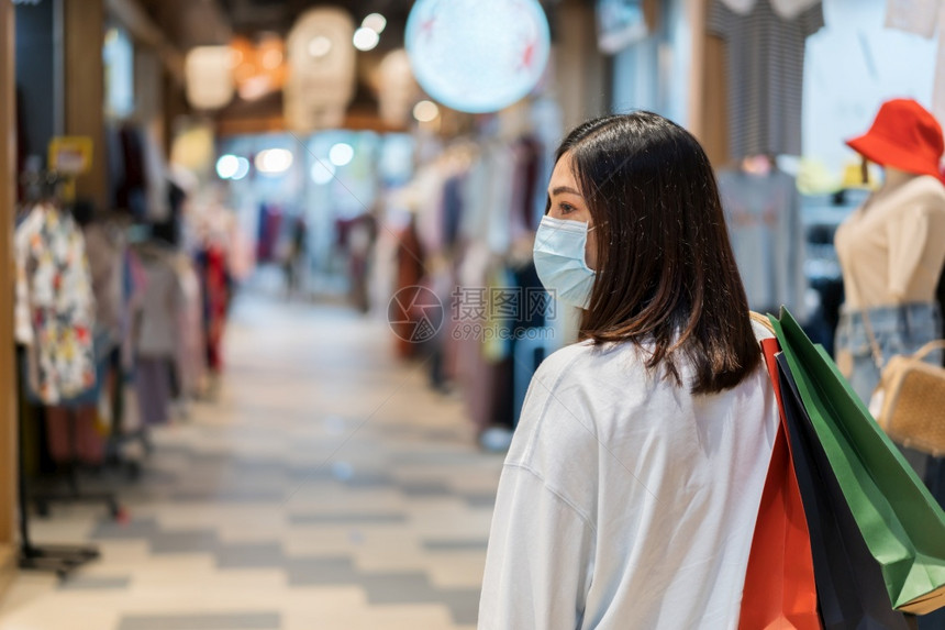 年轻妇女拿着购物袋在商场寻找衣服并戴医疗面具以防止科罗纳新冠大流行图片