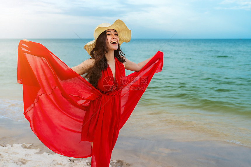 在海滨的风吹时手持红礼服的年轻女子图片