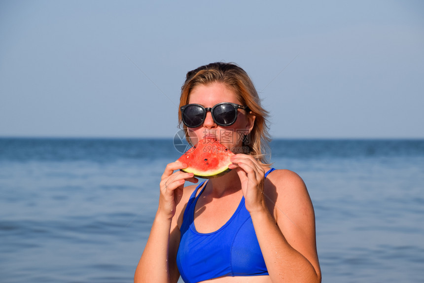 身着太阳镜的金发女郎在海边吃西瓜一只多汁的西瓜在一个女人的手中身着太阳眼镜的金发女郎在海边吃西瓜一只多汁的西瓜在一个女人的手中图片