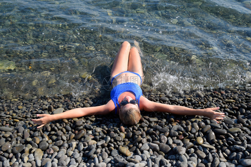一个穿蓝色比基尼的漂亮女孩躺在海滩上一半的水里落基海滩岸上的小比布透明海水蓝色比基尼的漂亮女孩一半在海滩的水里洛基海滩岸上的小比图片