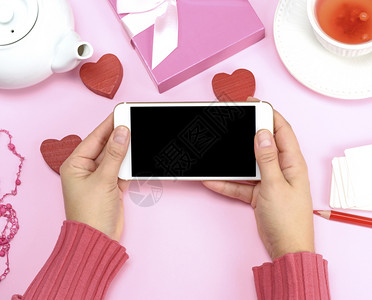 穿着粉红色毛衣的两只女手拿着白色智能手机空白黑屏顶视背景图片