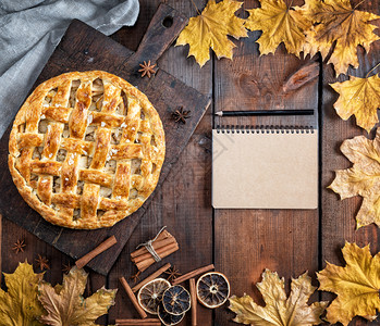 在棕色木板上烤整个圆苹果派泡面糕饼和一张空的纸条上面有食谱的棕色页面图片