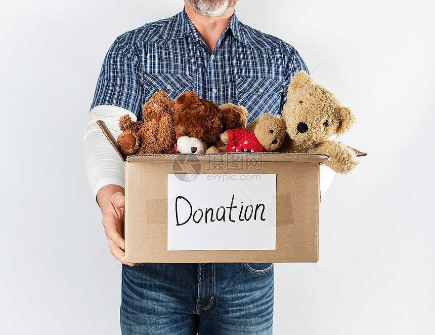 身穿蓝衬衫和牛仔裤的男子拿着一个大棕褐纸盒带儿童玩具帮助穷人的概念白种背景图片