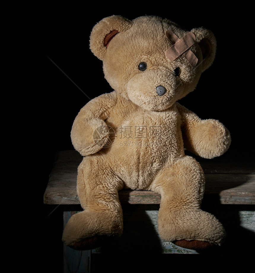 老棕褐色泰迪熊坐在木头表面石膏粘在前额上黑色背景图片