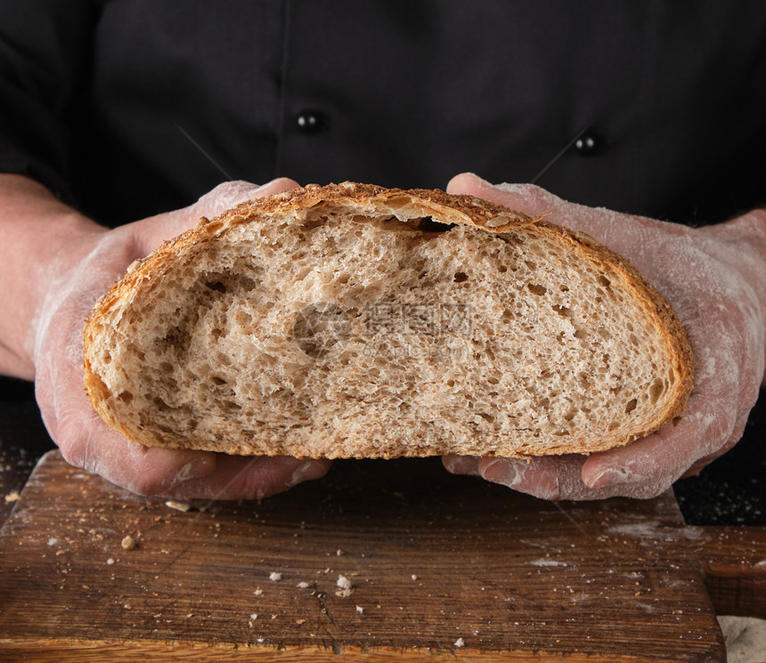身穿黑衣厨师服的手里握着一半用餐桌上的黑麦面粉烤碎掉结壳面包图片