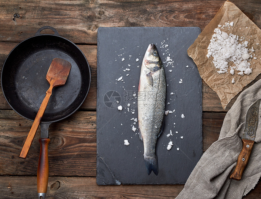 黑石墨板上的全新海贝斯鱼旁边是木制桌顶的空圆黑煎锅图片