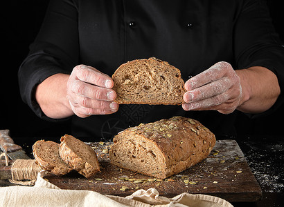 穿黑色制服的厨师继续切断一块面包用黑麦粉和南瓜种子烤成黑色背景图片