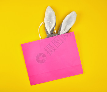 兔子从袋子里往外看黄色背景的白手柄长角粉红纸购物袋兔子耳朵从里伸出来平躺着背景