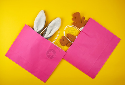 两袋粉红色纸用于购物放在一只兔子和黄色背景的鹿圣诞面罩内平铺图片