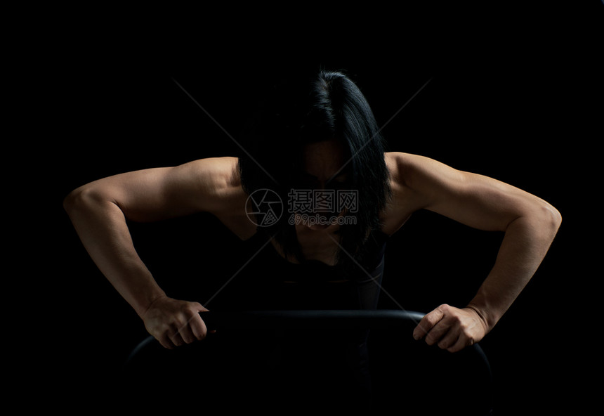 身穿黑发运动外貌的年轻女孩在黑暗背景下从表面推升运动者的肌肉很紧张图片