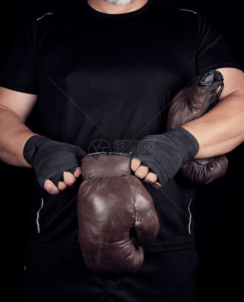 黑色衣服的成年肌肉男在比赛前双手戴上棕色皮衣拳击手套双被裹在黑色运动绷带中图片