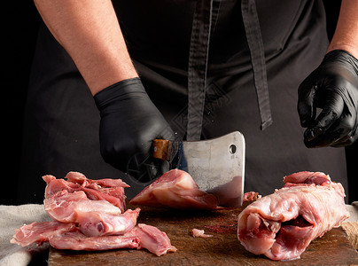 用黑色乳胶手套的厨师拿着一把大刀在棕色木制板上切碎生兔肉在黑暗背景下做饭背景图片
