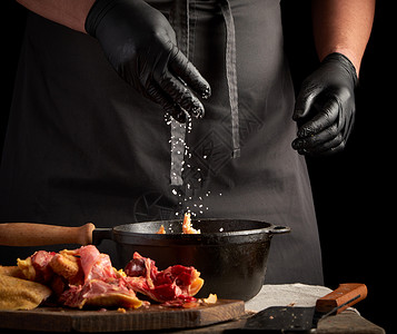 黑制服主厨和穿色制服的乳胶手套喷洒着白盐生鸡肉在黑铁煎锅里烤鸡肉做饭图片