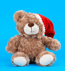 穿着红色圣诞礼帽的小棕色泰迪熊坐在蓝色背景上假日玩具图片