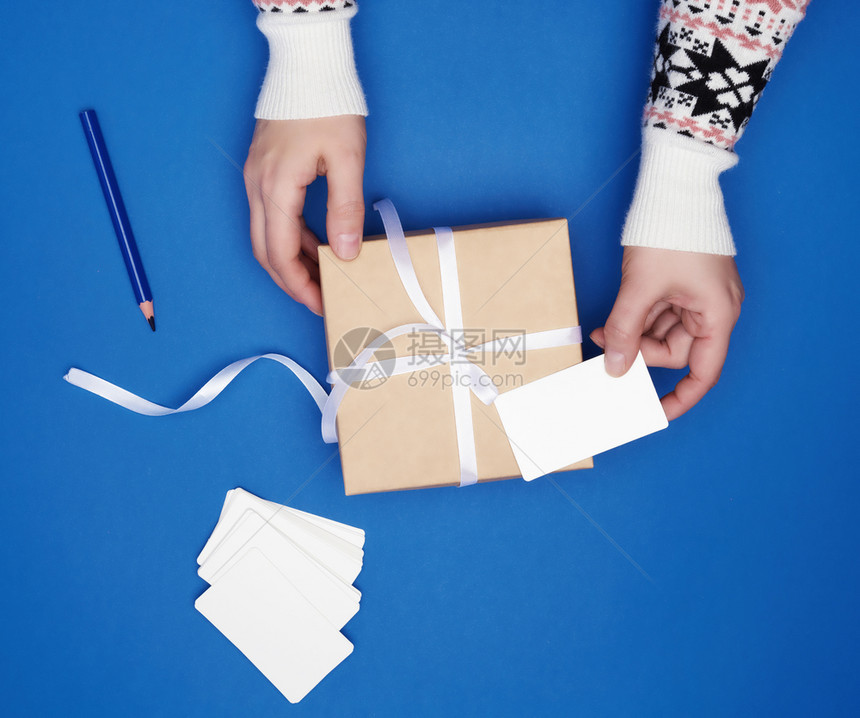 两只女手拿着一个纸板礼品箱用白色丝带和空纸牌捆绑在深蓝背景最美经典趋势颜色上图片