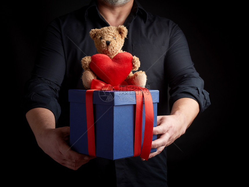 身穿黑衬衫的成年男子拿着一个带红色丝的蓝方盒子坐在棕色泰迪熊顶上图片