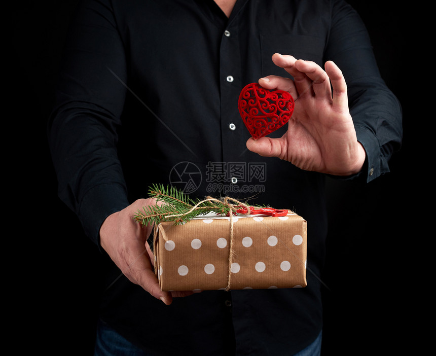 成人男子身穿黑衬衫站在暗背景上手拿着棕纸包的礼物左手拿着红雕刻的装饰心这是假日问候的概念图片