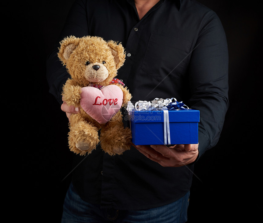 身穿黑衬衫的成人男子拿着一个蓝色方格盒子上面绑着白色丝带和棕泰迪熊心胸如2月14日情人节的祝贺概念图片