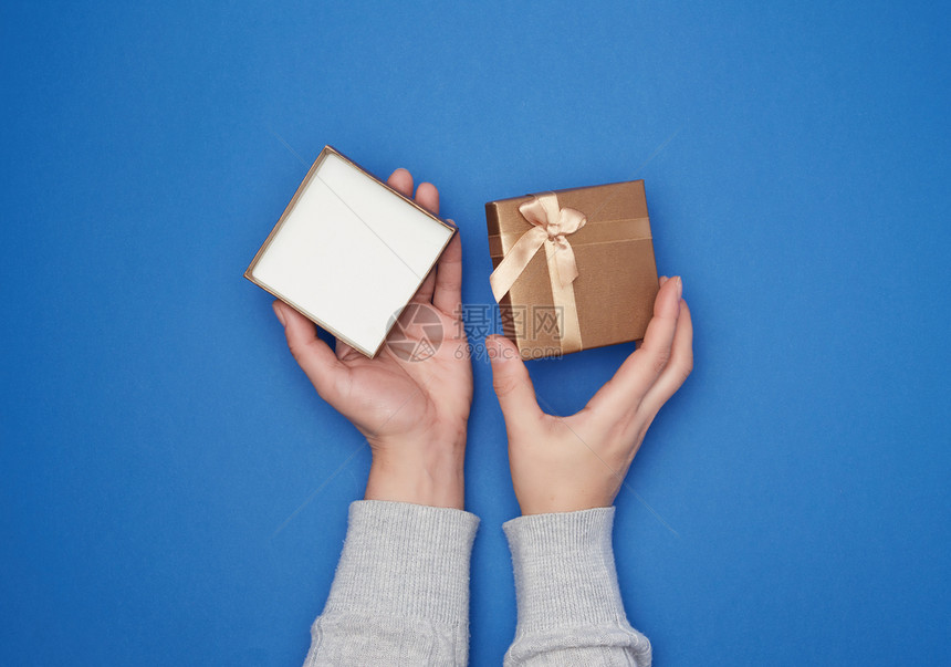 灰色毛衣中的女手握着开的纸板礼品盒用蓝色背景趋势颜假日背景的弓头挂在纸板上图片