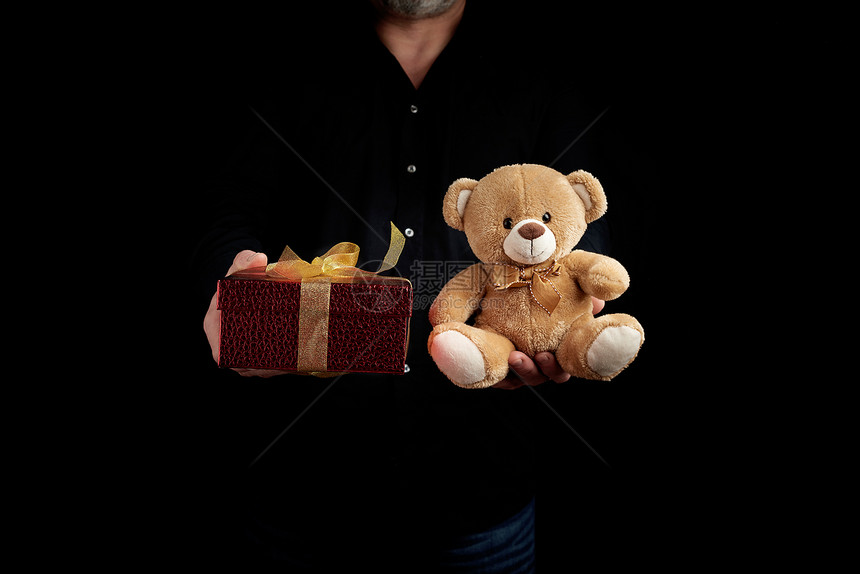 一名身穿黑色衬衫的成年男子手持一个用金色丝带和棕色泰迪熊绑着的红色方形盒子低调地构思着2月14日情人节的祝贺图片