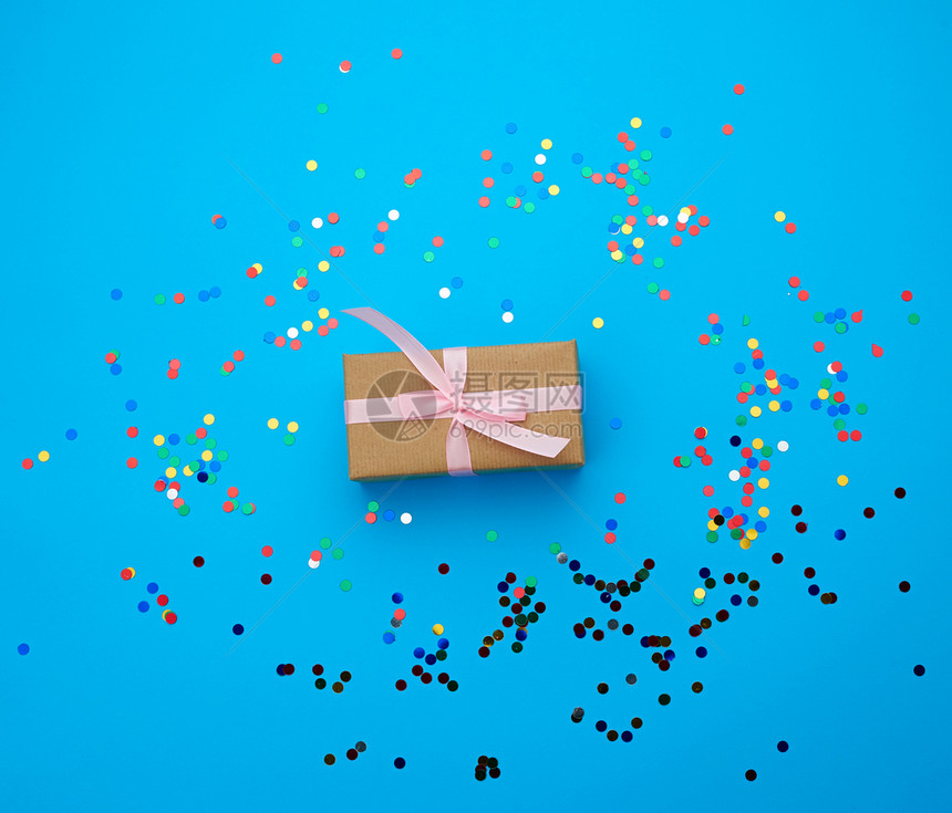 棕色礼盒用纸包裹用丝绸粉色丝带系在蓝色背景上配以多色闪亮的彩色纸屑作为生日情人节的节日背景图片