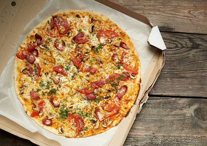 用熏香肠蘑菇西红柿奶酪和薄饼烤圆披萨在木制餐桌上的开板纸盒中食物被切成片顶视背景图片