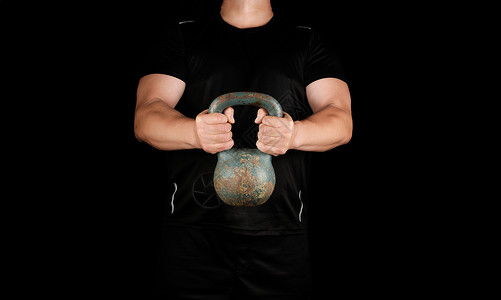 身穿黑色衣服的成年强健运动员在伸展的手臂上用黑色背景握着铁叮当背景图片