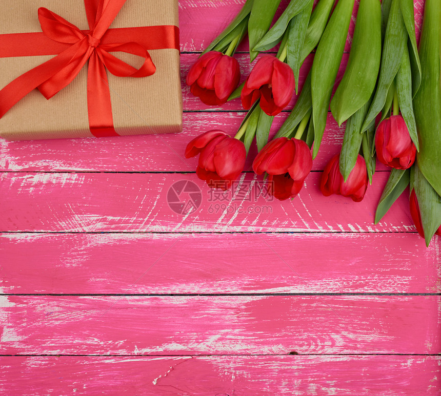 红色盛开的郁金香花束配有绿色叶子棕手纸包着的礼物以及粉红色木质背景的丝绸红色带图片