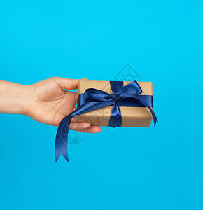 手握着一个盒子礼物包在棕色的Kraft纸上绑在蓝色背景的丝绸带上赠送礼物的概念背景图片