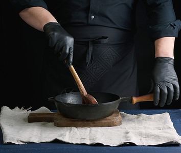 黑制服和乳胶手套的男烹饪者用黑色制服和乳胶手套用黑色的柄和底黑铸铁锅盘将老式木勺子食品搅拌成木勺子背景图片
