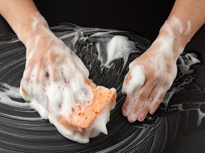 黑色海绵白色肥皂泡沫中的两个女手握着一湿海绵擦黑表面清洁和洗表面的概念顶视背景