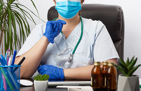 身穿制服和蓝色乳胶手套的医生坐在办公室工作桌前在诊所接待病人的概念图片