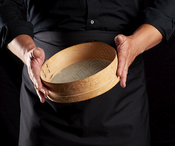 身穿黑色制服的男子拿着空的老古董圆木筛子来选面粉厨师站在黑色背景上关图片