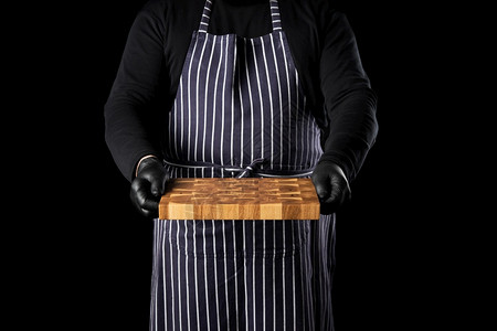 身穿条纹蓝色围裙和黑衣服的男主厨站在黑色背景的对面手里握着一个长方形木制厨房切削板背景图片