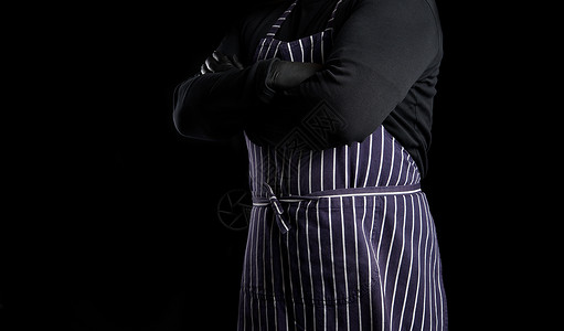 身穿条纹蓝色围裙和黑衣服的男厨背着黑色景胸口交叉他的手臂复制空间图片