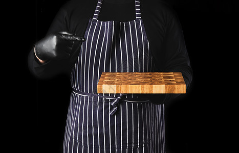 男黑衣服素材身穿条纹蓝色围裙和黑衣服的男主厨站在黑色背景的对面手里握着一个长方形木制厨房切开板关上背景