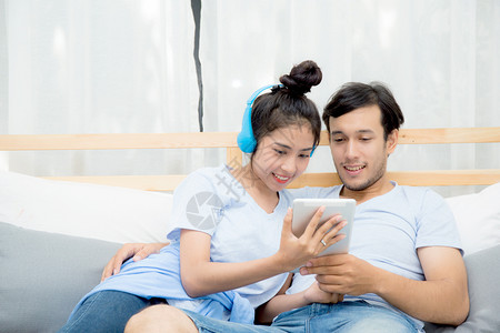 美丽的亚洲年轻夫妇听着床上有平板电脑的音乐爱约会年轻夫妇坐在床上用平板电脑坐在床上背景图片