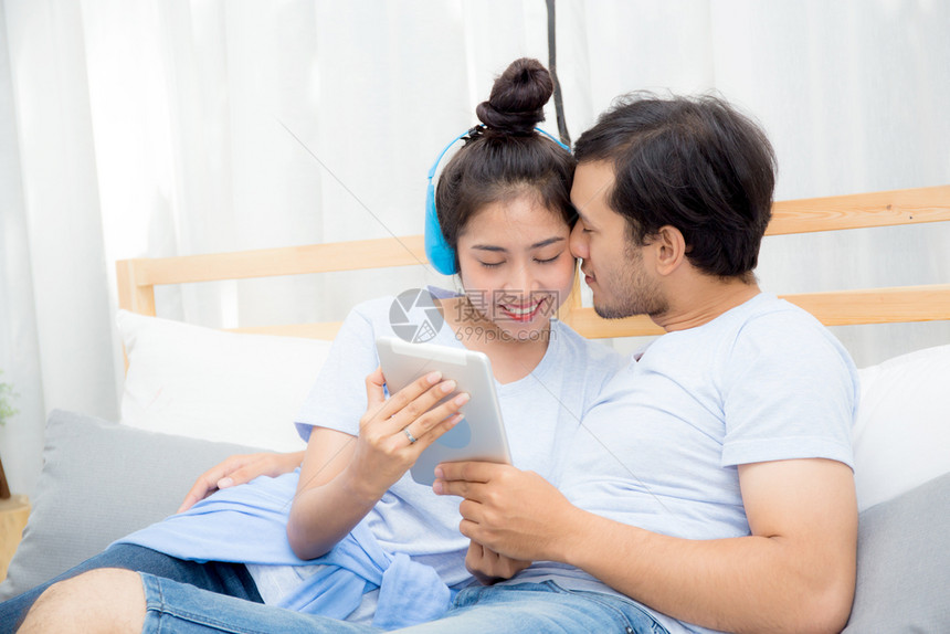 美丽的亚洲年轻夫妇听着床上有平板电脑的音乐爱约会年轻夫妇坐在床上用平板电脑坐在床上图片
