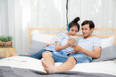 美丽的亚洲年轻夫妇听着床上有平板电脑的音乐爱约会年轻夫妇坐在床上用平板电脑坐在床上背景图片