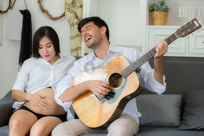 丈夫弹吉他美丽的怀孕妈在沙发的客厅里家人和放松概念图片