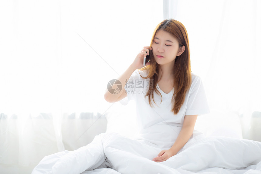 美丽的年轻女士早上在床微笑坐放松女孩用移动智能电话在卧室聊天交流概念图片
