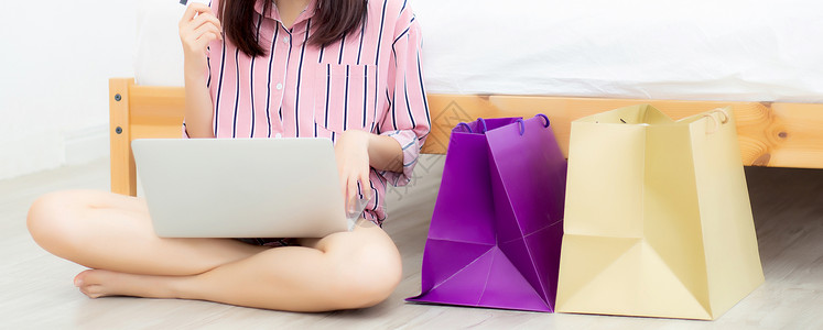 Banner网站美貌的亚洲妇女在线购物手持笔记本电脑坐在房间里的地板上持有信用卡购买和物袋的女孩以及生活方式概念背景图片