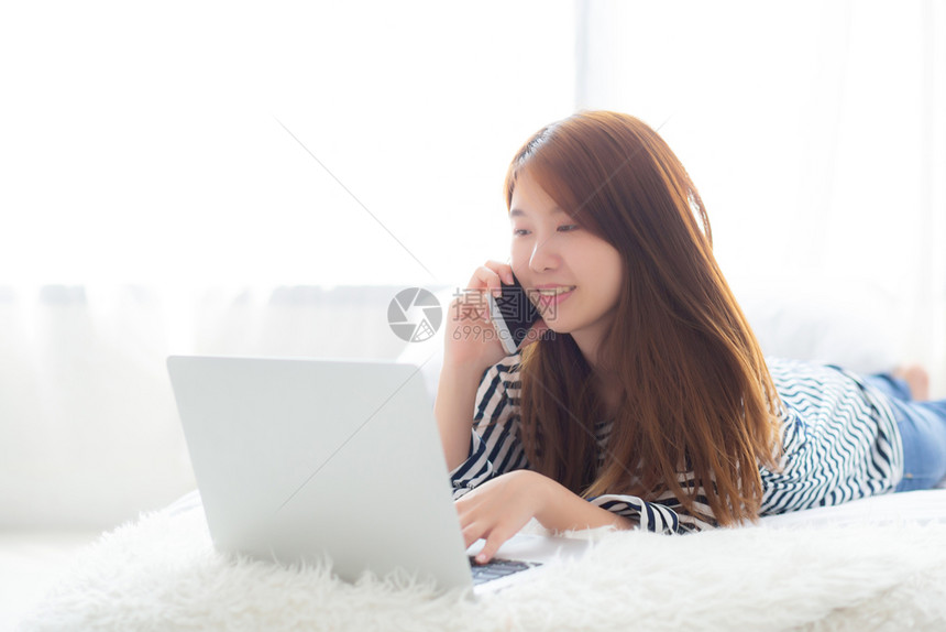 躺在床上的美丽亚洲年轻女使用笔记本电脑和在卧室讲手机智能电话休闲和放松自由职业图片