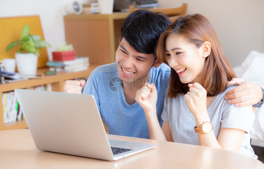 年轻和的亚裔夫妇在用笔记本电脑做时带着微笑和快乐坐在卧室里使用兴奋和满意的笔记本电脑商业和成功的概念图片
