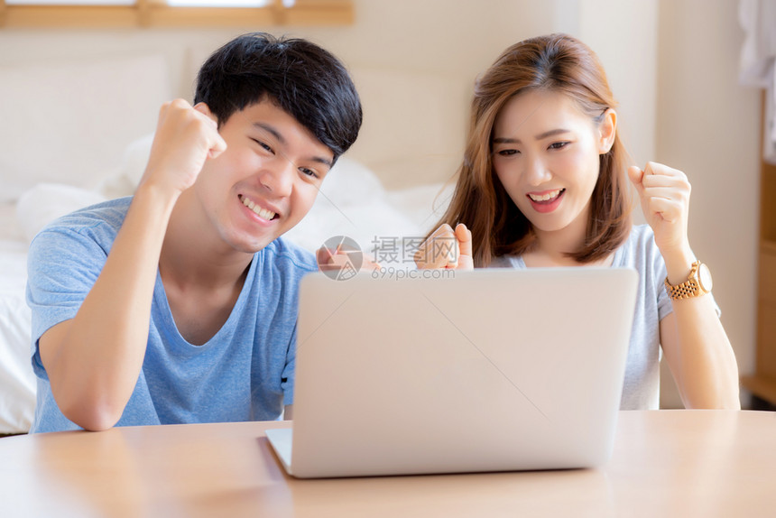 年轻和的亚裔夫妇在用笔记本电脑做时带着微笑和快乐坐在卧室里使用兴奋和满意的笔记本电脑商业和成功的概念图片