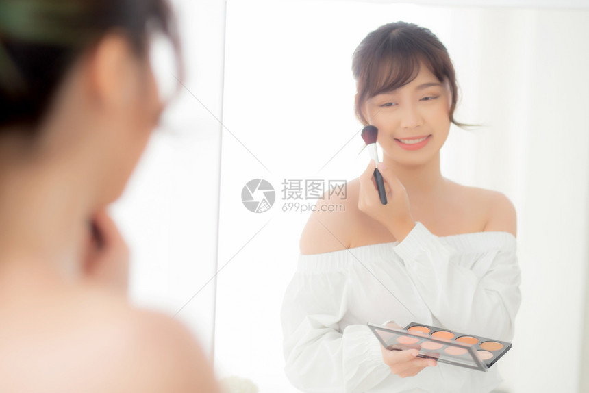 美容肖像年轻的亚洲女微笑着面容的镜子在卧室里化妆时用刷脸美丽的女孩拿着脸红皮肤照护化妆桌时装概念图片