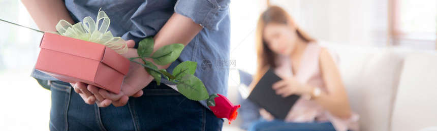 美丽的年轻夫妇男人藏着红色礼物盒和玫瑰花在家里惊喜丈夫和妻子快乐情人节或生日结婚家庭爱的概念横幅网站图片